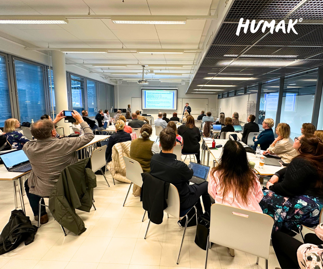 Luokkahuone on täynnä ammattikorkeakoulujen asintuntijoita ja rehtoreita. He kuuntelevat tarkasti esitystä, joka pidetään luokkahuoneessa. Kankaalle on heijastettu PowerPoint-esitys. Jotkut ottavat kännykän kameralla kuvaa esityksestä.
