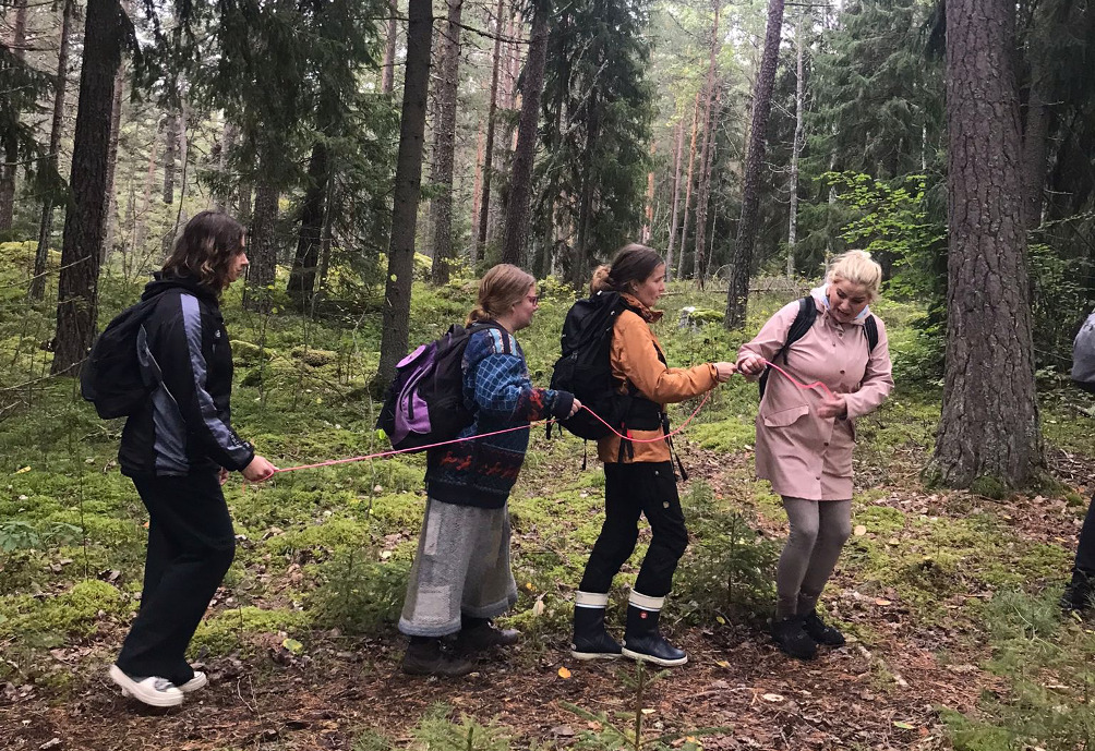 Ohjaaja pitää metsässä narun päästä kiinni ja vetää perässään kolmea narussa kiinni olevaa ihmistä, joilla on silmät kiinni