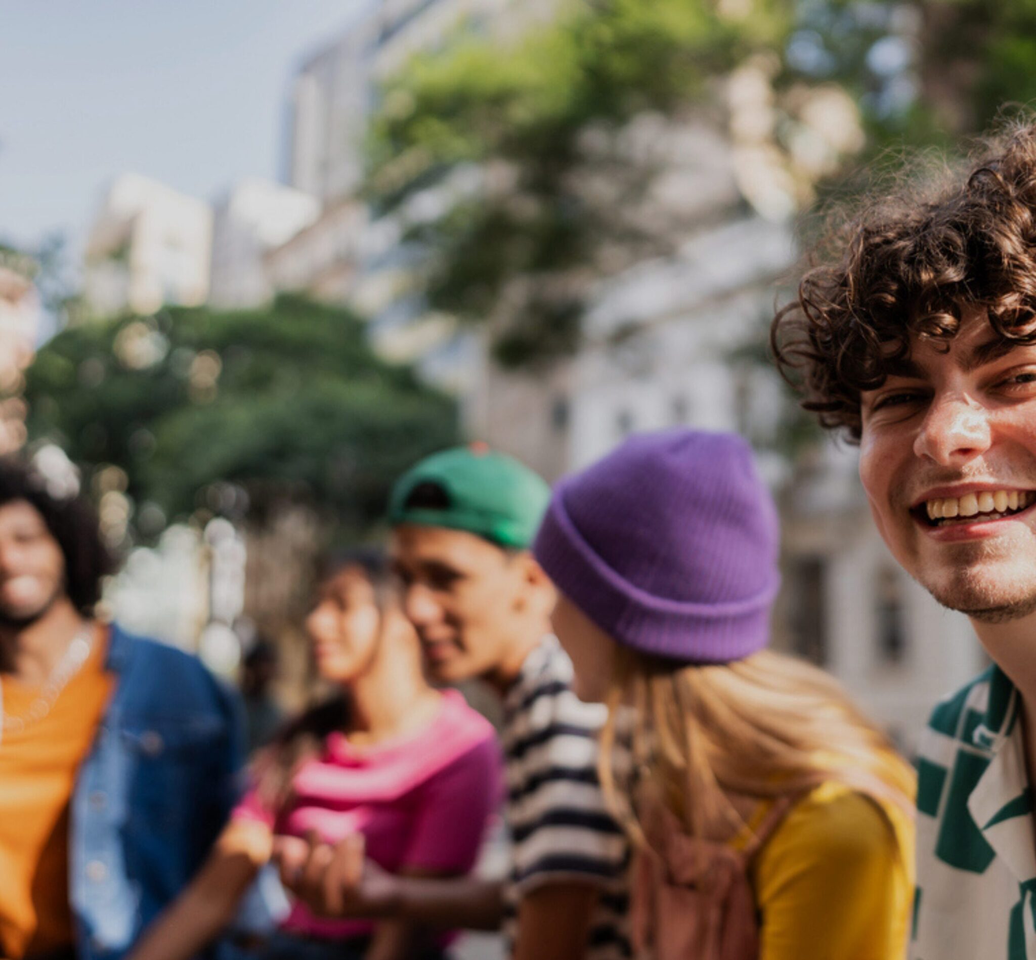 oukko nuoria istuu ulkona kaupunkimaisemassa aurinkoisena päivänä. He keskustelevat rennosti penkillä, ja etualalla oleva henkilö käyttää vihreä‑valkoista kuviollista paitaa. Taustalla näkyy puita ja rakennuksia.