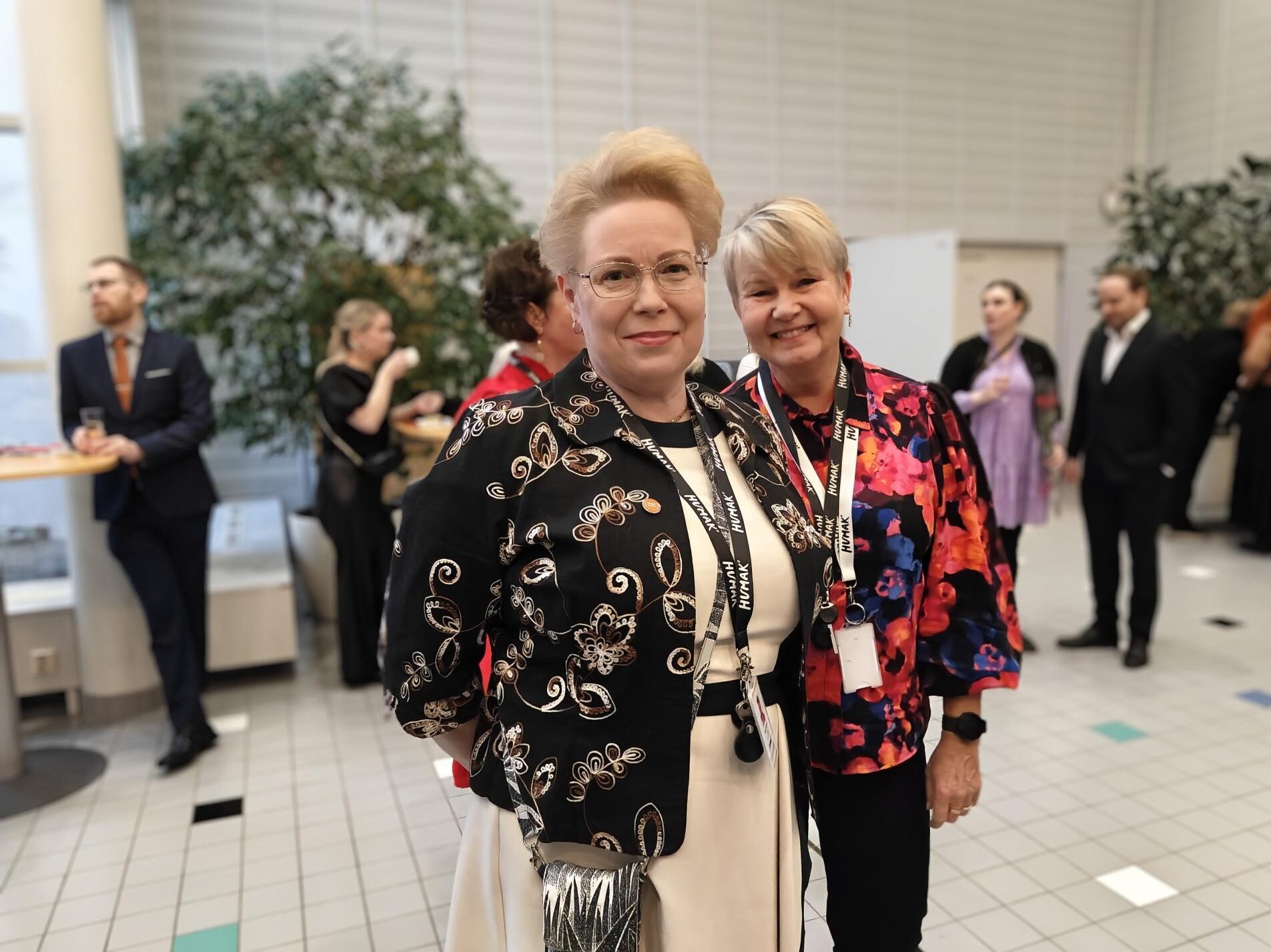 Kaksi naista kuvassa, etualla oleva hymyilee suu kiinni, okapään takana oleva hymyilee hampaat näkyen. Lievästi taustahimmennystä käytetty taustan osalta, jossa muutamia henkilöitä seisoo aulatilassa.