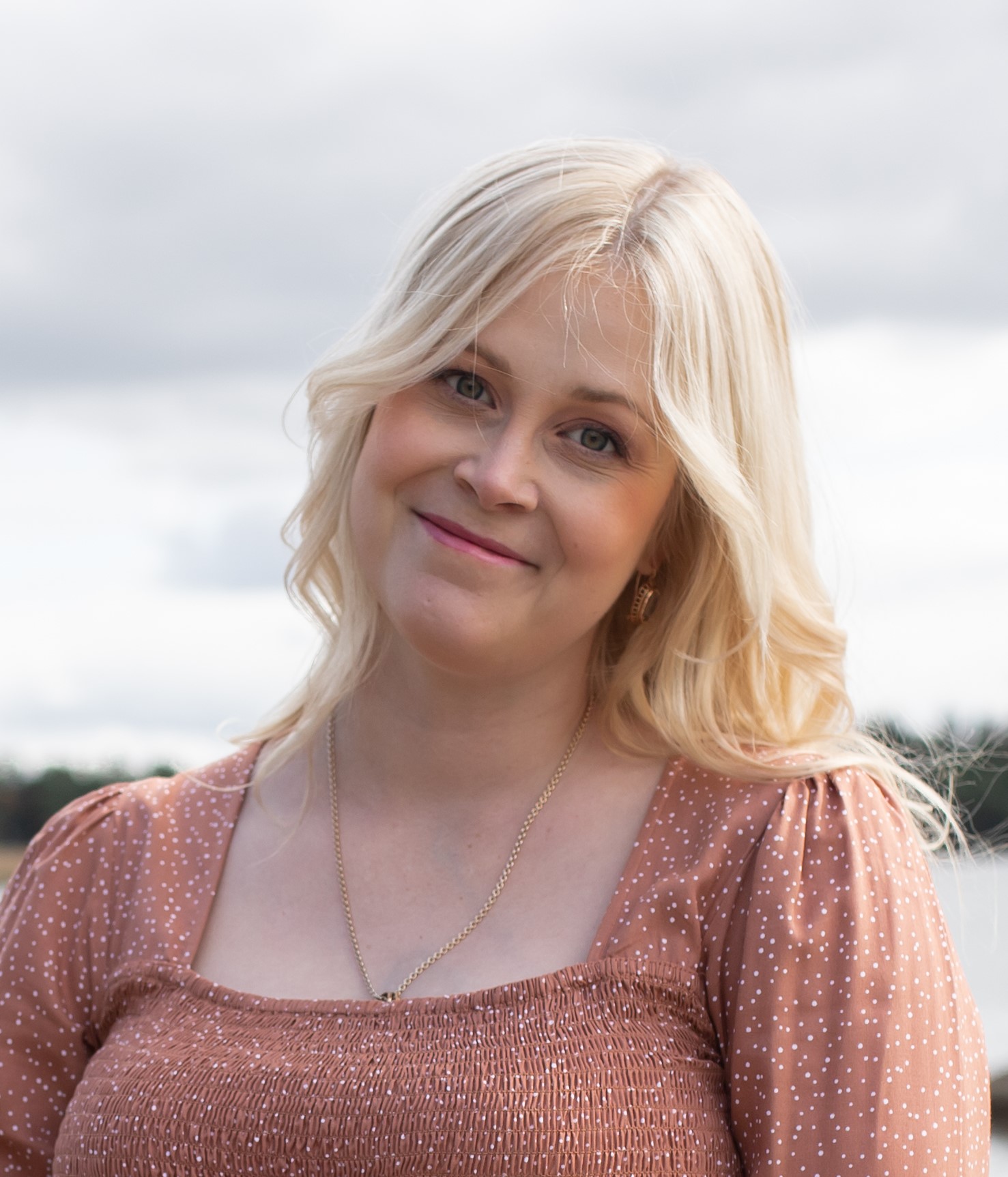 Vaalehiuksinen nainen hymyilee vienosti kameralle, taustalla vesimaisema.