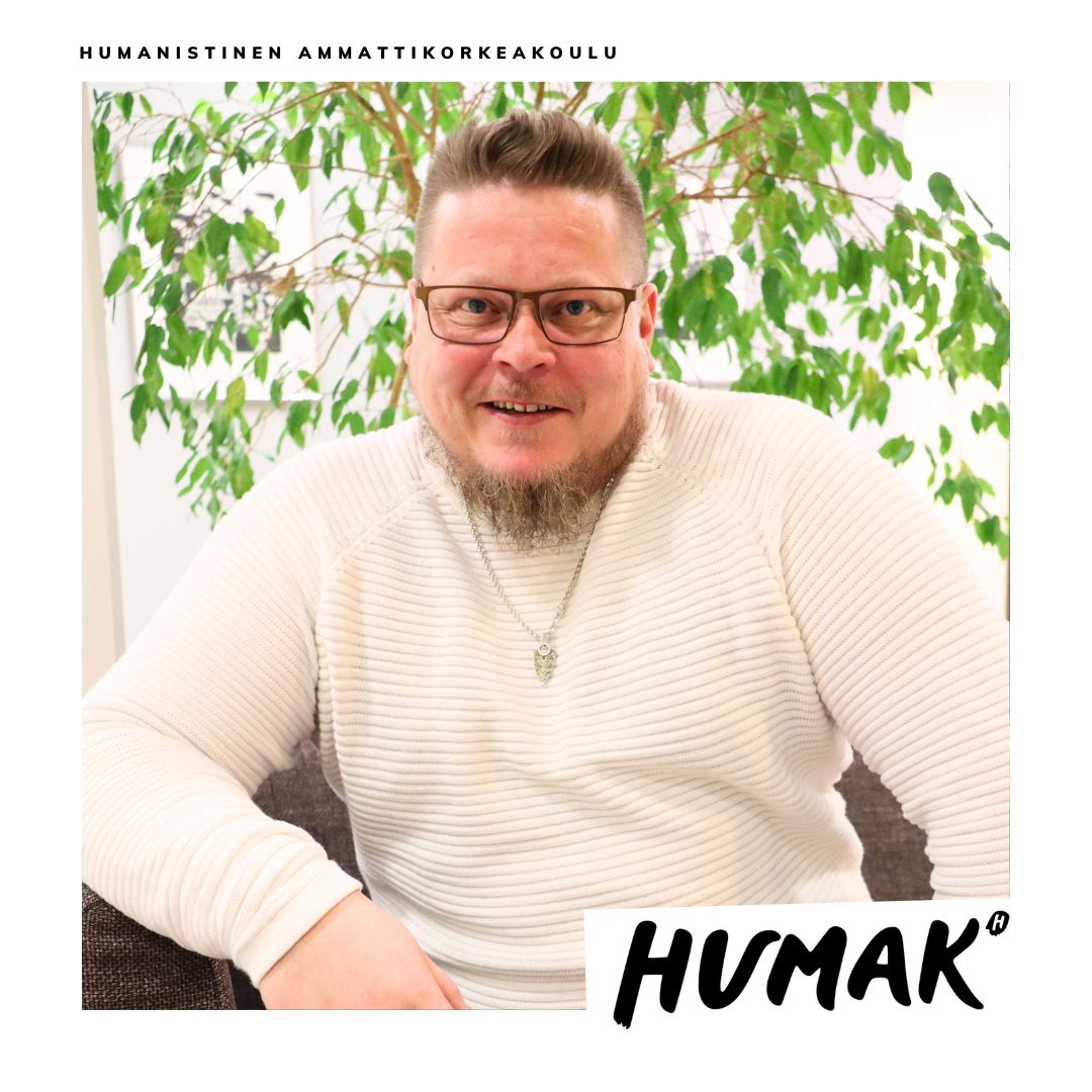 Vaalea mies katsoo hymyillen kohti kameraa. Miehellä on vaaleat lyhyet hiukset, silmälasit ja vaalea parta sekä päällään valkea paita. Takana on viherkasveja