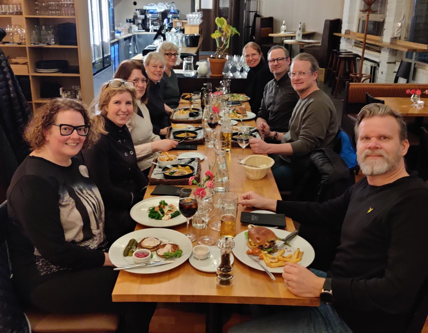 Nio leende personer sitter vid restaurang bord i trä med maten serverad på tallrikar.