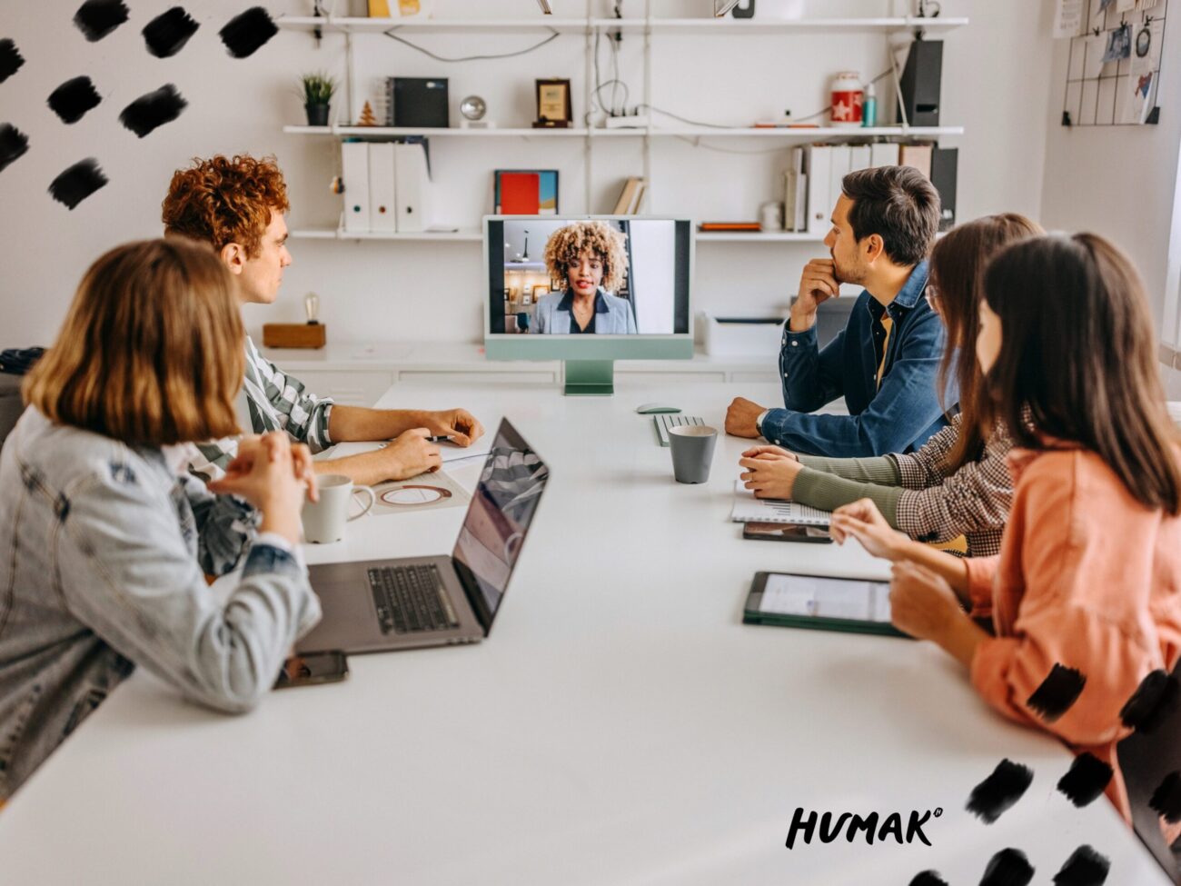 Ryhmä ihmisiä istuu neuvottelupöydän ääressä ja seuraa videokokousta tietokoneen näytöltä modernissa toimistotilassa.