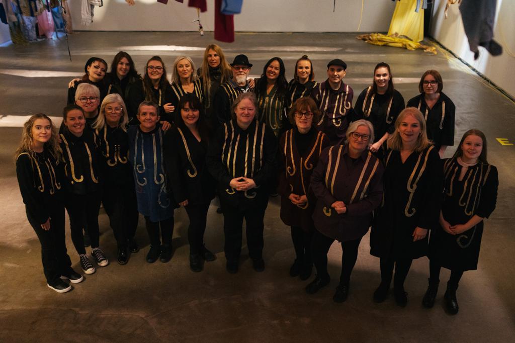 Yli 20 ihmistä seisoo salissa mustissa vaatteissa ja katsoo kameraan. 