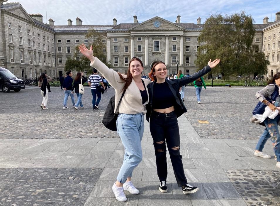 Kaksi henkilöä poseeraa iloisesti kädet ylhäällä suuren tiilisen rakennuksen edustalla.