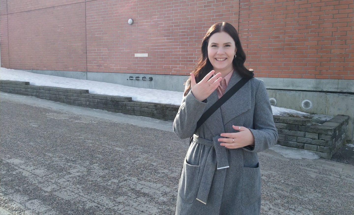 Pitkähiuksinen nainen pitkässä takissa, olkalaukun hihna vartalon päällä, kädet viittovat. Taustalla tiiliseinä, ulkokuva.