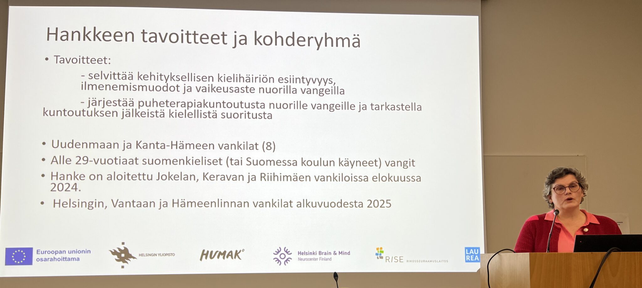 Luennoitsija seisoo valkokankaan edessä. Diassa näkyy hankkeen tavoitteet ja kohderyhmä.