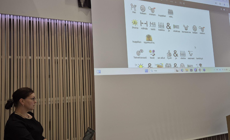 kuvan vasemmassa alakulmassa nainen. Kuvan oikealla puolella valkokankaalle heijastettu kuvakirjoitusteksti. 