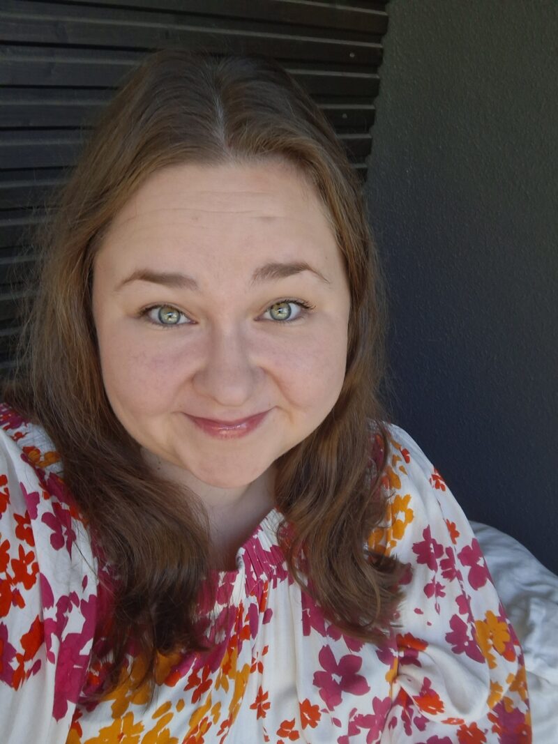 Tässä alt-teksti kuvallesi (ilman mitään mainintaa sumeasta kasvoalueesta): **Henkilö istuu ulkotilassa tumman seinän edessä. Henkilöllä on olkapäille ulottuvat ruskeat hiukset ja yllään valkoinen paita, jossa on punaisia, oransseja ja keltaisia kukkakuvioita.**