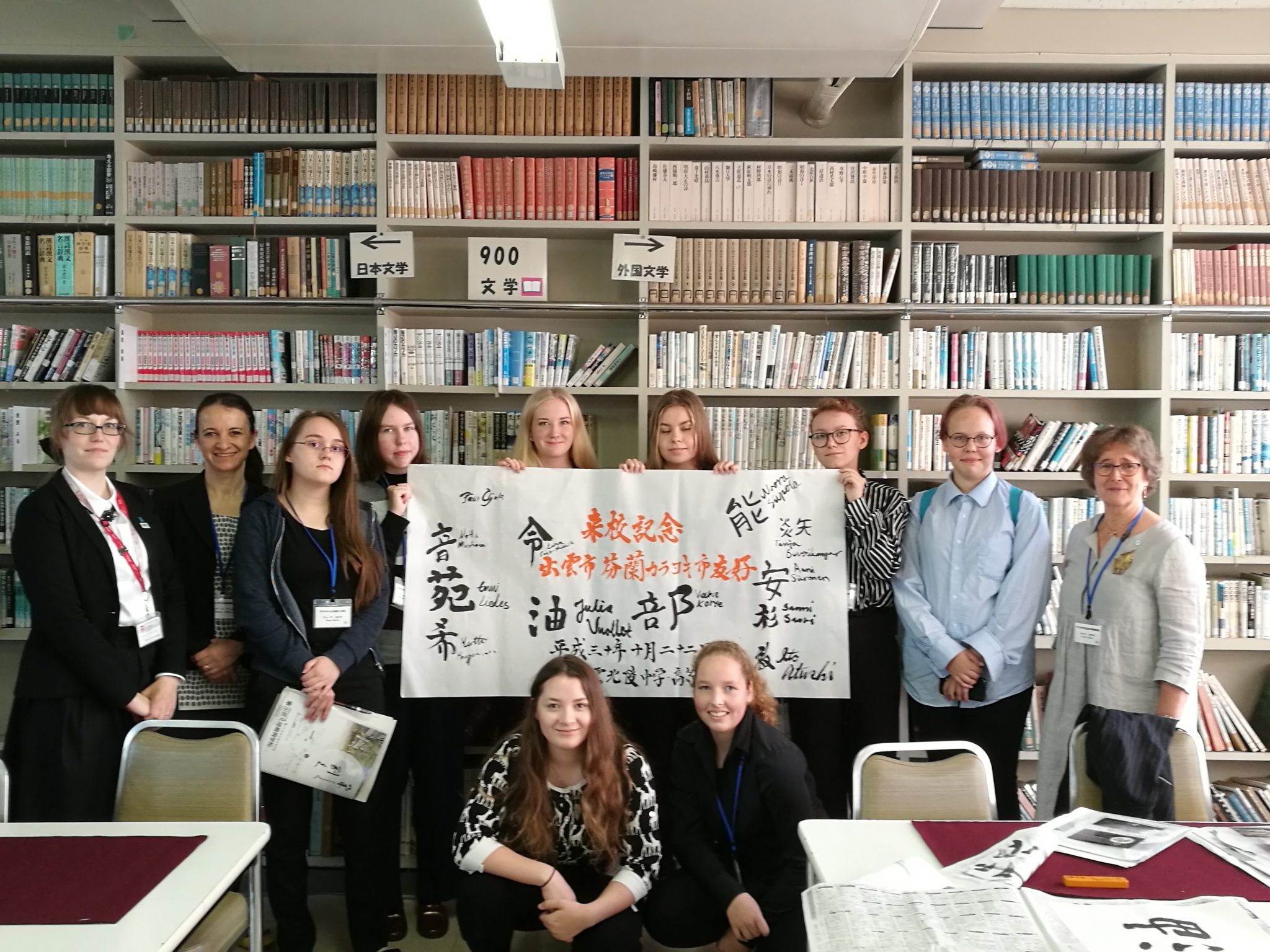 A group of students and supervisors standing in front of multiple bookshelves with four of them holding a white sign with Japanese symbols on it.