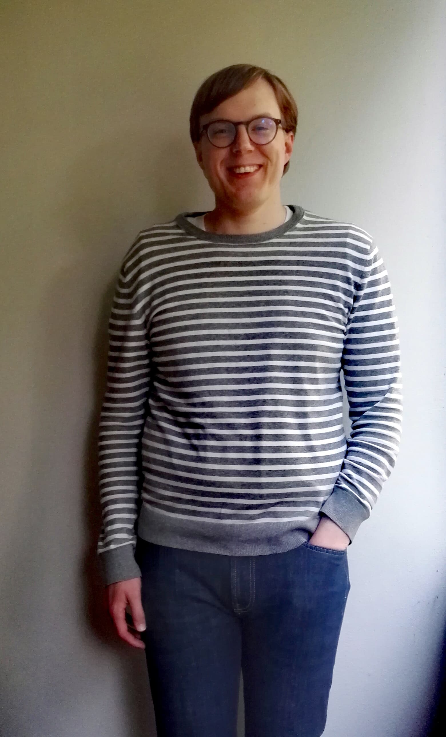 Raitapaitainen mies seisoo valkoista seinää vasten hymyillen, vasen käsi farkkujen taskussa.