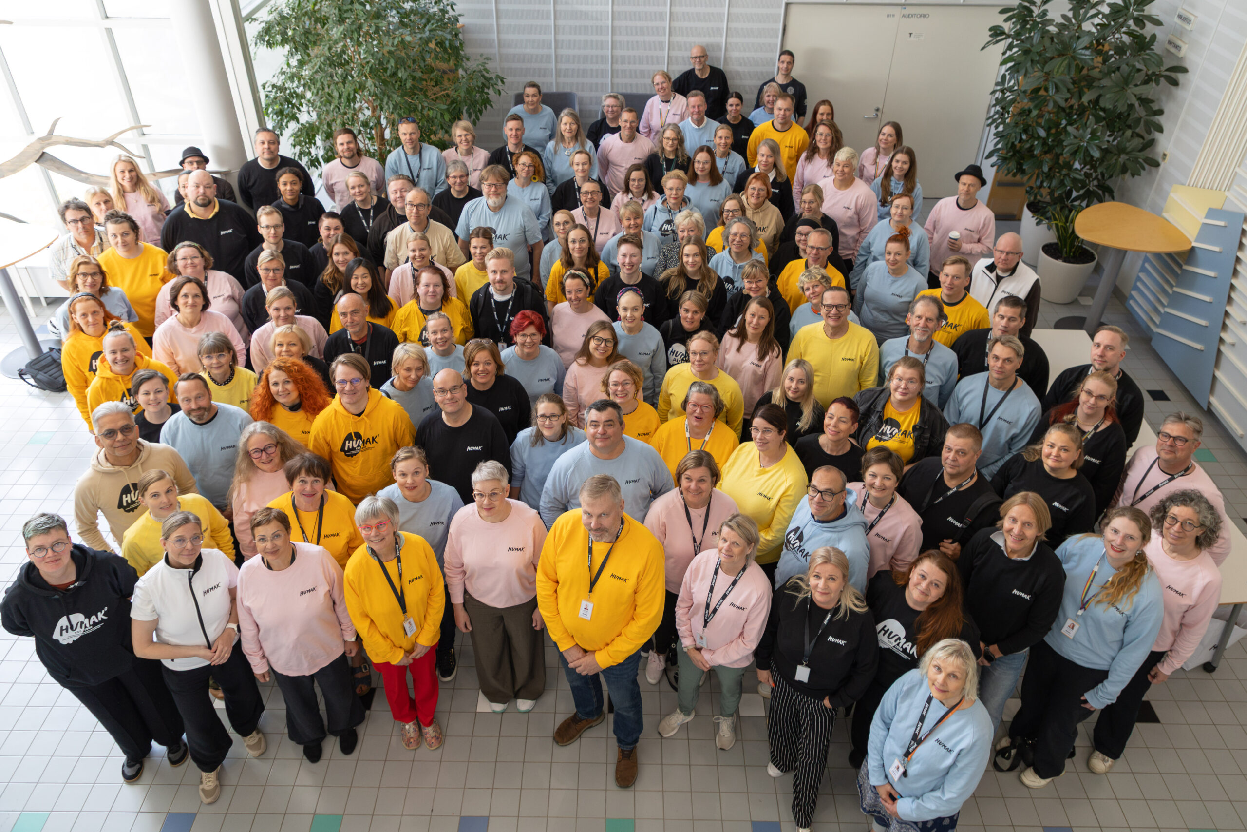 Group portrait of about 100–120 staff members standing closely together in an indoor atrium, photographed from above, most smiling and facing the camera while wearing a mix of yellow, pink, light blue and dark tops with Humak logo.