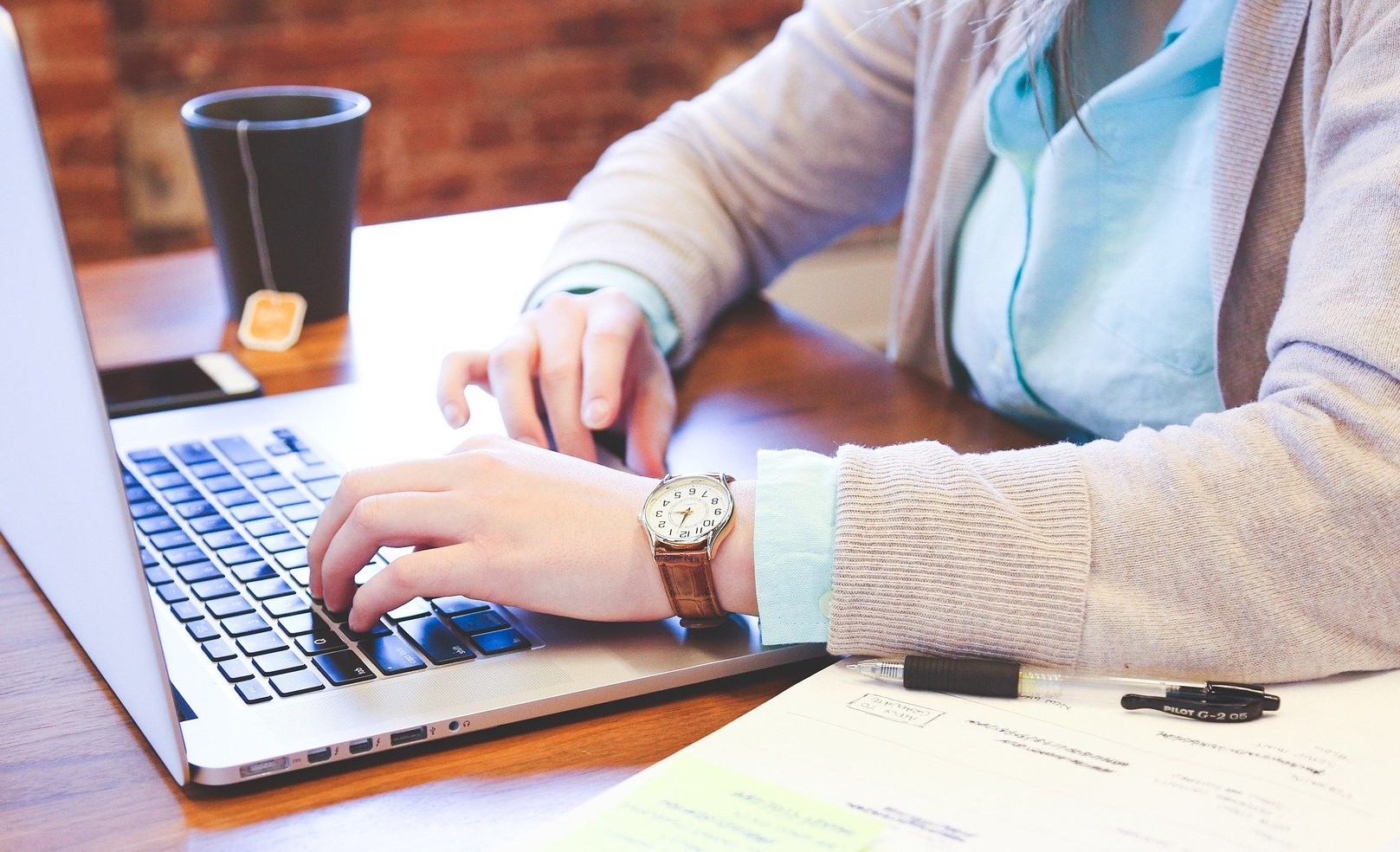 Kuvassa kannettavan tietokoneen näppäimistö, jolla henkilön kädet, vasemmassa kädessä kello. Henkilöllä vaaleansininen pitkähihainen paita, jonka päällä beige neuletakki. Pöydällä on kuppi, josta roikkuu teepussin naru.