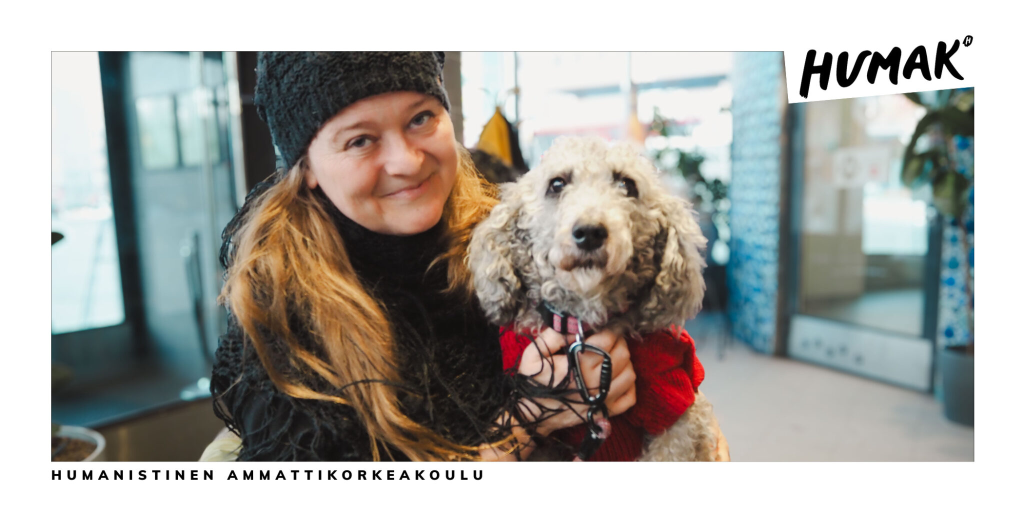 Mustiin pukeutunut pitkähiuksinen nainen katsoo hymyillen kameraan. Hänellä on sylissään vaalea villakoira. Taustalla näkyy valoisa ravintolan aula. 