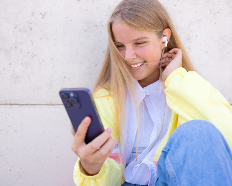 Nuori henkilö istuu ulkona seinää vasten, pitää älypuhelinta kädessään ja kuuntelee kuulokkeita värikkääseen takkiin pukeutuneena.
