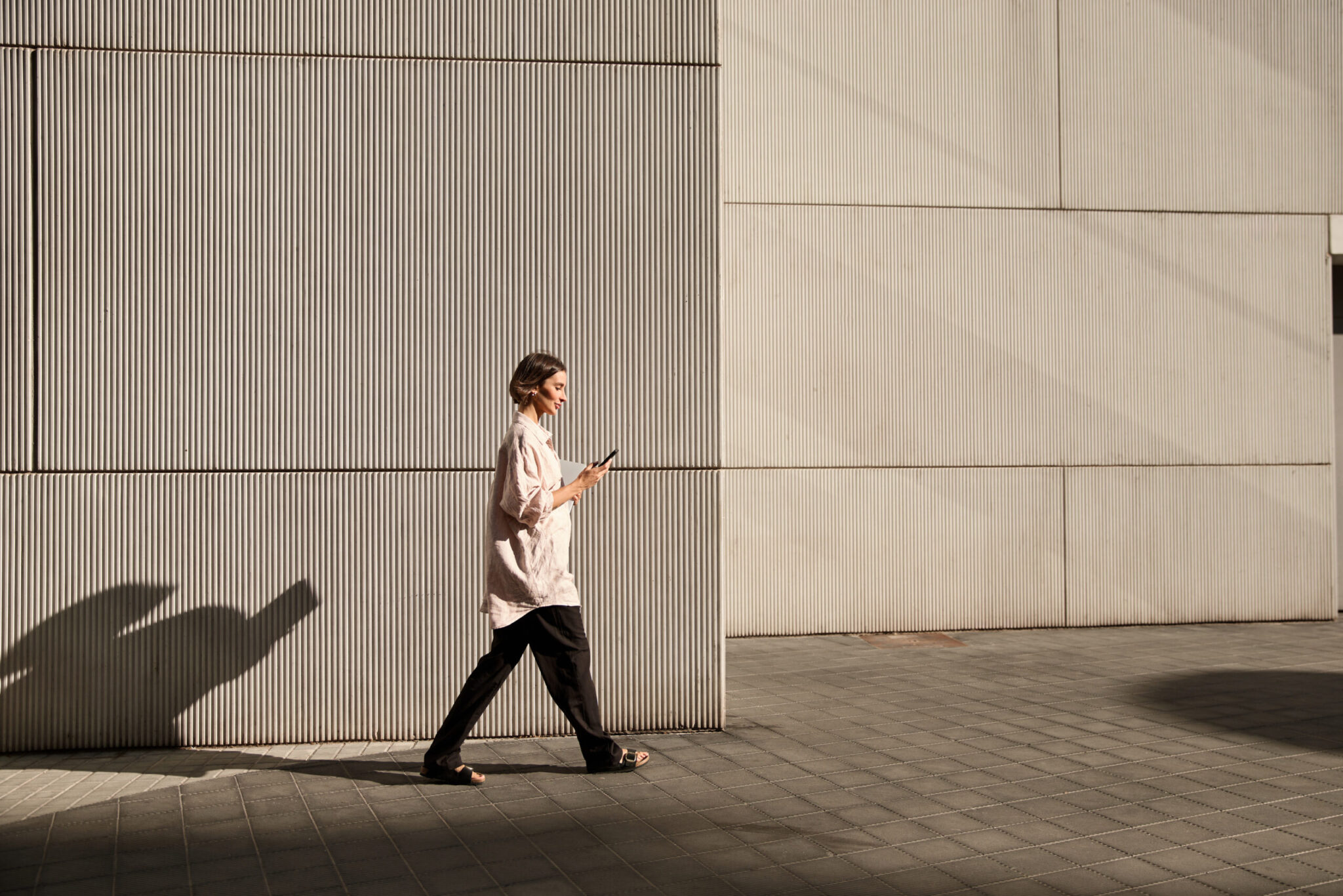 Henkilö kävelee modernin, vaalean rakennuksen seinän vieressä ja katsoo älypuhelintaan; iltapäivän aurinko luo pitkän varjon katukiveykselle.