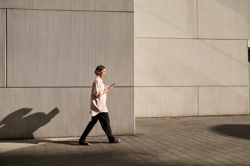 Henkilö kävelee modernin, vaalean rakennuksen seinän vieressä ja katsoo älypuhelintaan; iltapäivän aurinko luo pitkän varjon katukiveykselle.