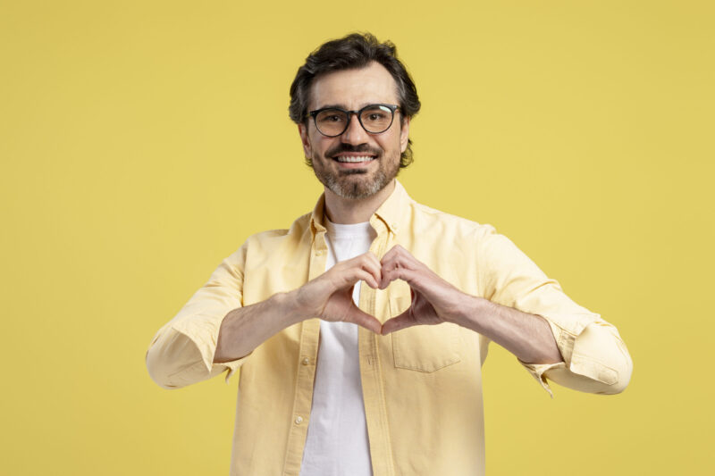 Smiling man creating a heart shape with his hands, radiating love, care, and affection against a vibrant yellow background, capturing positive emotions and human connection. Sign language concept