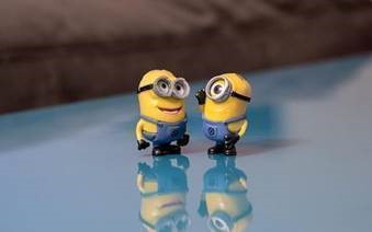Two characters from the animated film Minions are smiling and having a conversation. The other yellow character has one eye and the other has two eyes. They are both wearing strong glasses, blue overalls and black boots. The background is faded.