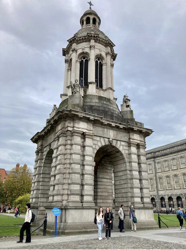 Kuvassa on noin 30 metriä korkea kellotorni, joka on pääasiassa rakennettu harmaasta graniitista ja muodostuu kolmesta osasta. Alin osa on neliön muotoinen. Neliön kulmat muodostuvat pilareista, joiden välissä on jokaisella sivulla holvikaaret, joista pääsee kulkemaan kellotornin alitse. Pilareiden päällä, jokaisessa kulmassa on patsaat. Kellotornin keskimmäinen ja ylin osa ovat lieriön muotoiset. Keskimmäinen osa on isompi kuin ylin osa. Lieriön muotoisten osien seinät muodostuvat kapeista holvikaarista. Kellotornin katolla on risti. Kellotornin seinissä ja katoissa on kuvioita ja kaiverruksia. Kellotornin edustalla seisoo ihmisiä ja sen takana näkyy nurmikenttää, puita sekä vanhoja rakennuksia.