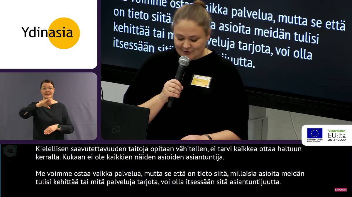 Kuvankaappus seminaarin verkkolähetyksestä. Vasemmalla on kaksi ruutua, joissa ylemmässä on Ydinasia-hankkeen logo ja alapuolella on viittomakielen tulkkaus. Oikealla on Tanja puhumassa mikrofoniin. Alareunassa näkyy kirjoitustulkkaus.