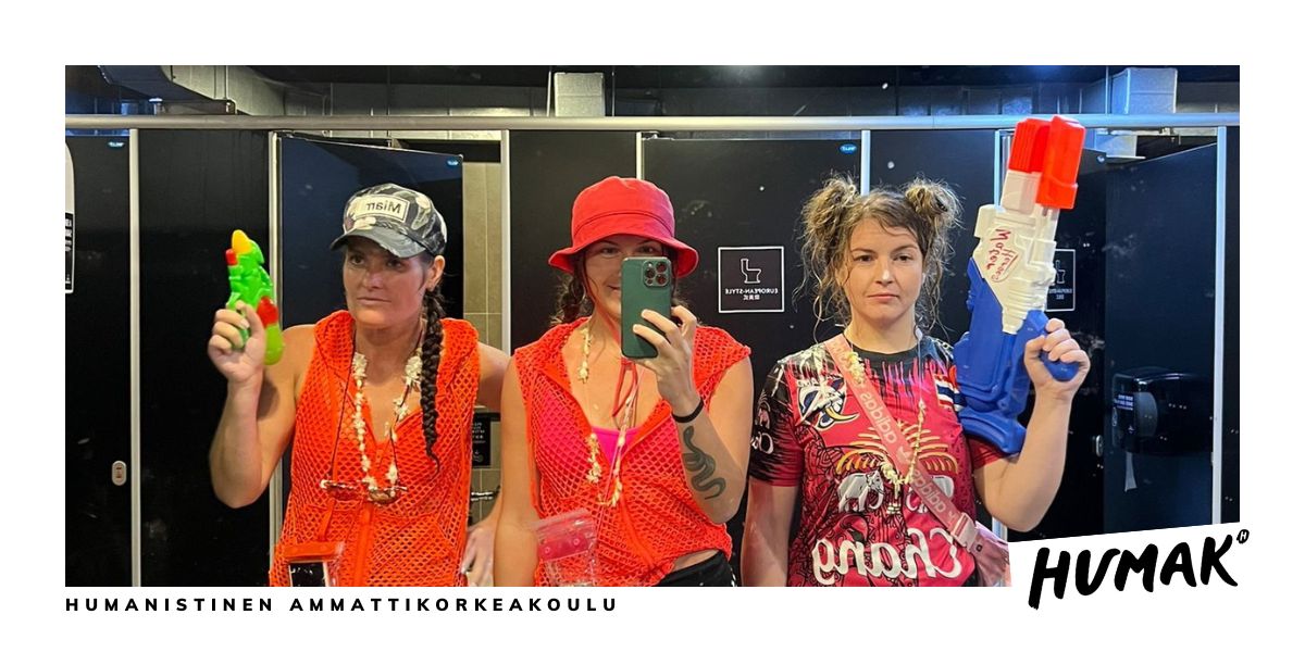 Kolme naista seisoo vierekkäin punaisissa vaatteissa vesipyssyt kädessä. Keskimmäinen ottaa kolmikosta peiliselfien. 