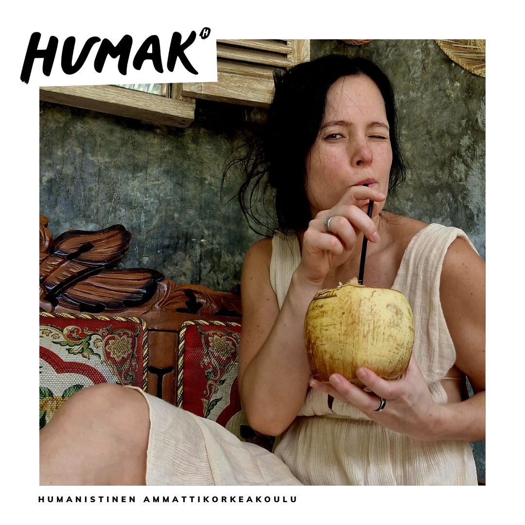 Tummahiuksinen, vaaleaan kesämekkoon pukeutunut nainen juo pillillä kookospähkinästä ja katsoo silmää iskien kameraan. 