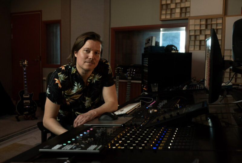 Tumma hiuksinen mies istuu studiopöydän äärellä, jossa miksereitä.