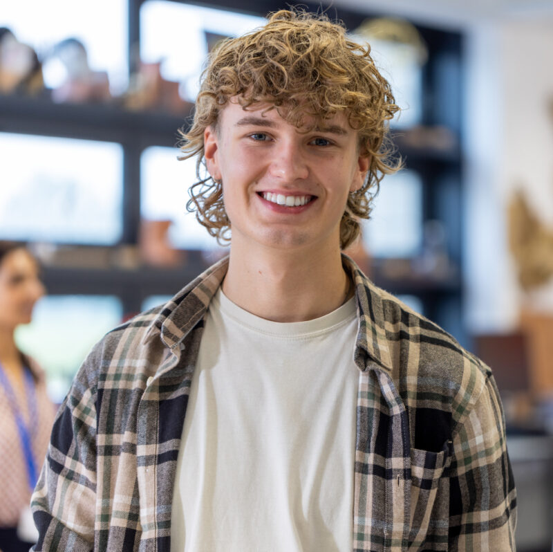 Luokkahuoneessa seisova henkilö rennossa asennossa, päällään ruudullinen flanellipaita ja vaalea t-paita. Taustalla näkyy oppimisympäristön kalusteita sekä muita ihmisiä työskentelemässä tai keskustelemassa.