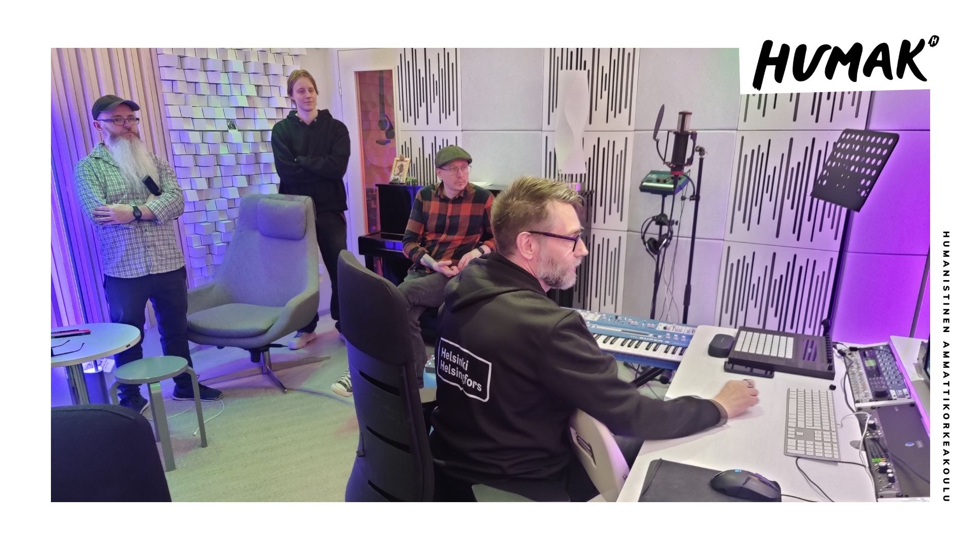 Kuvassa on vaalea moderni äänitysstudio. Helsingin kaupungin huppariin pukeutunut mies istuu pöydän ääressä ja käyttää äänisuunnittelulaitteita, ja kolme muuta miestä katselee. 
