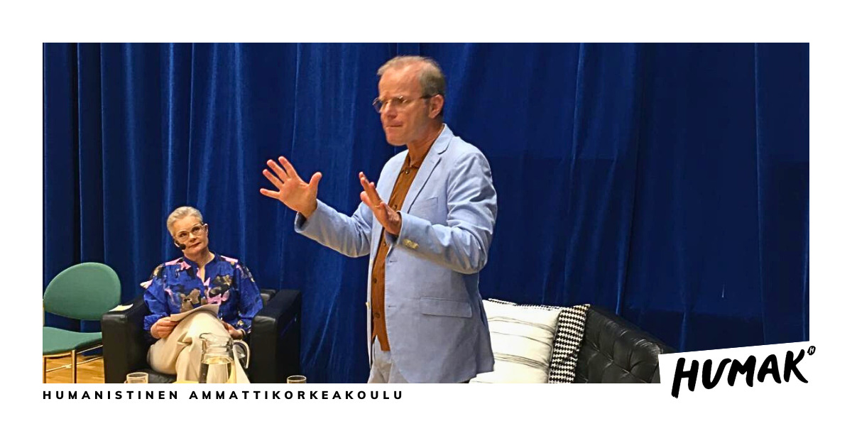 Siniseen pukuun pukeutunut mies seisoo ja viittoo sohvan edessä. Lyhythiuksinen nainen istuu tuolilla ja tarkastelee. Taustalla on paksu, tummansininen verho. 