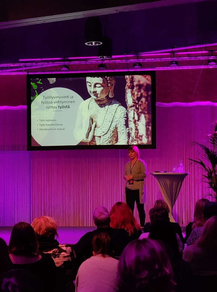 Työhyvinvoinnin seminaarissa otettu kuva, jossa puhumassa vaaleahiuksinen nainen.
