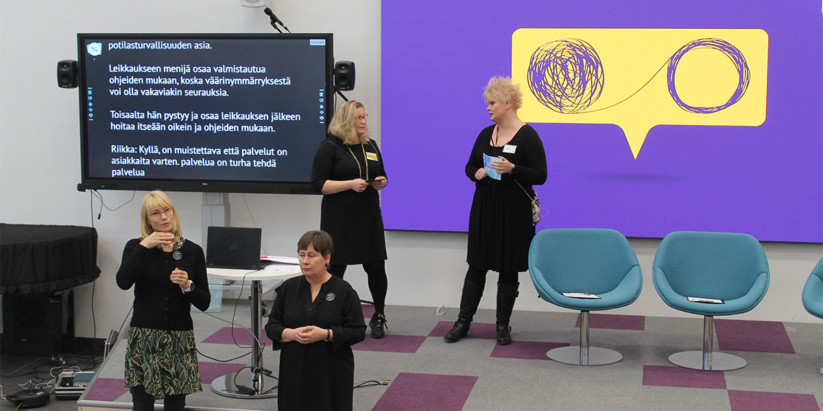 Kuvassa Soilimaria ja Riikka seisovat lavalla. Vasemmalla puolella on kirjoitustulkkauksen ruutu ja heidän takanaan Ydinasia-hankkeen logo. Kaksi viittomakielen tulkkia seisoo lavan edessä.