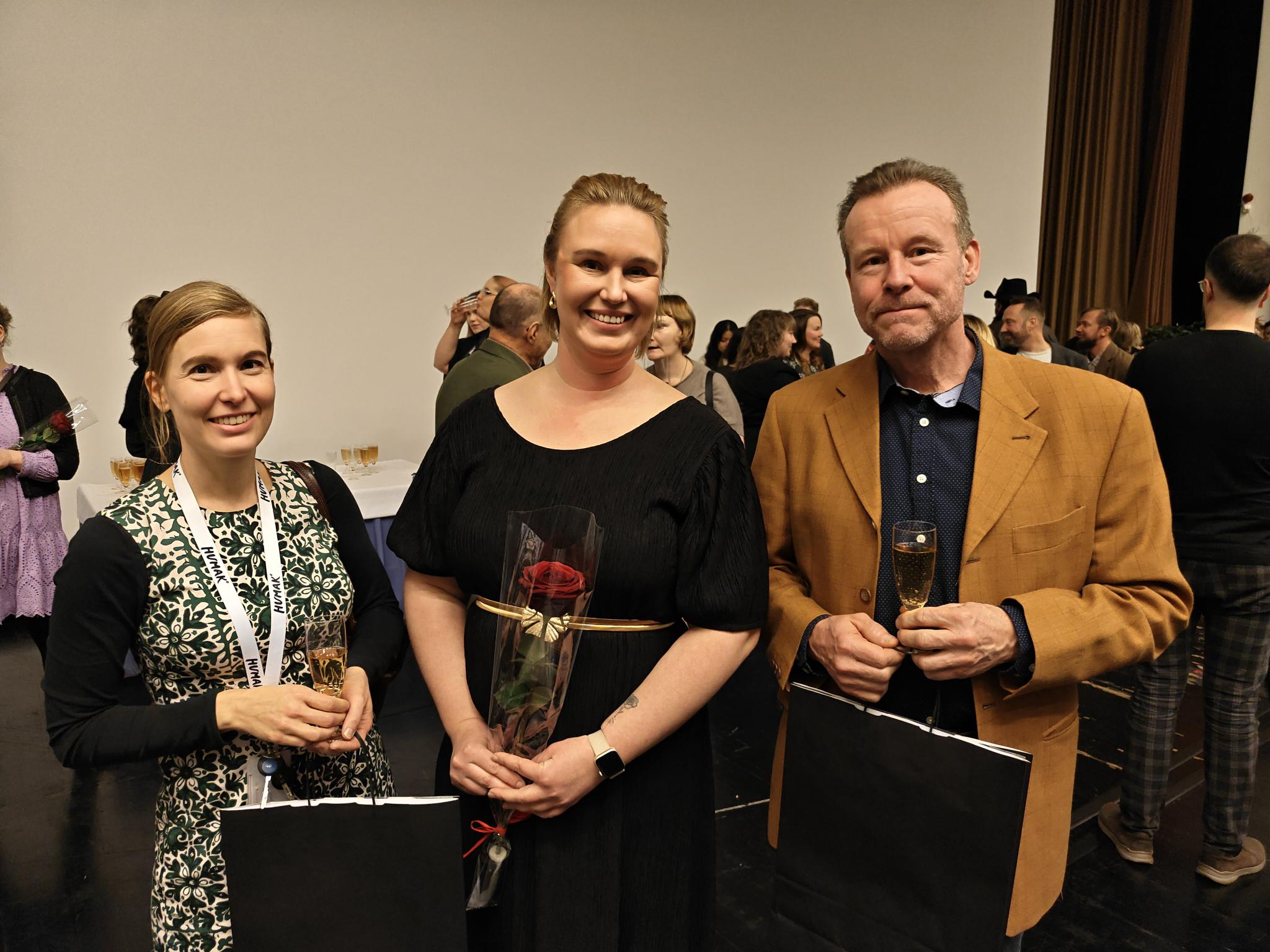 Kolme aikuista hymyilee kameralle juhlatilaisuudessa. He seisovat vierekkäin sisätilassa muiden vieraiden keskellä, kädessään kuohuviinilasit ja mustat lahjakassit; keskimmäinen henkilö pitää kädessään punaista ruusua. Taustalla näkyy lisää ihmisiä ja tarjoilupöytä.
