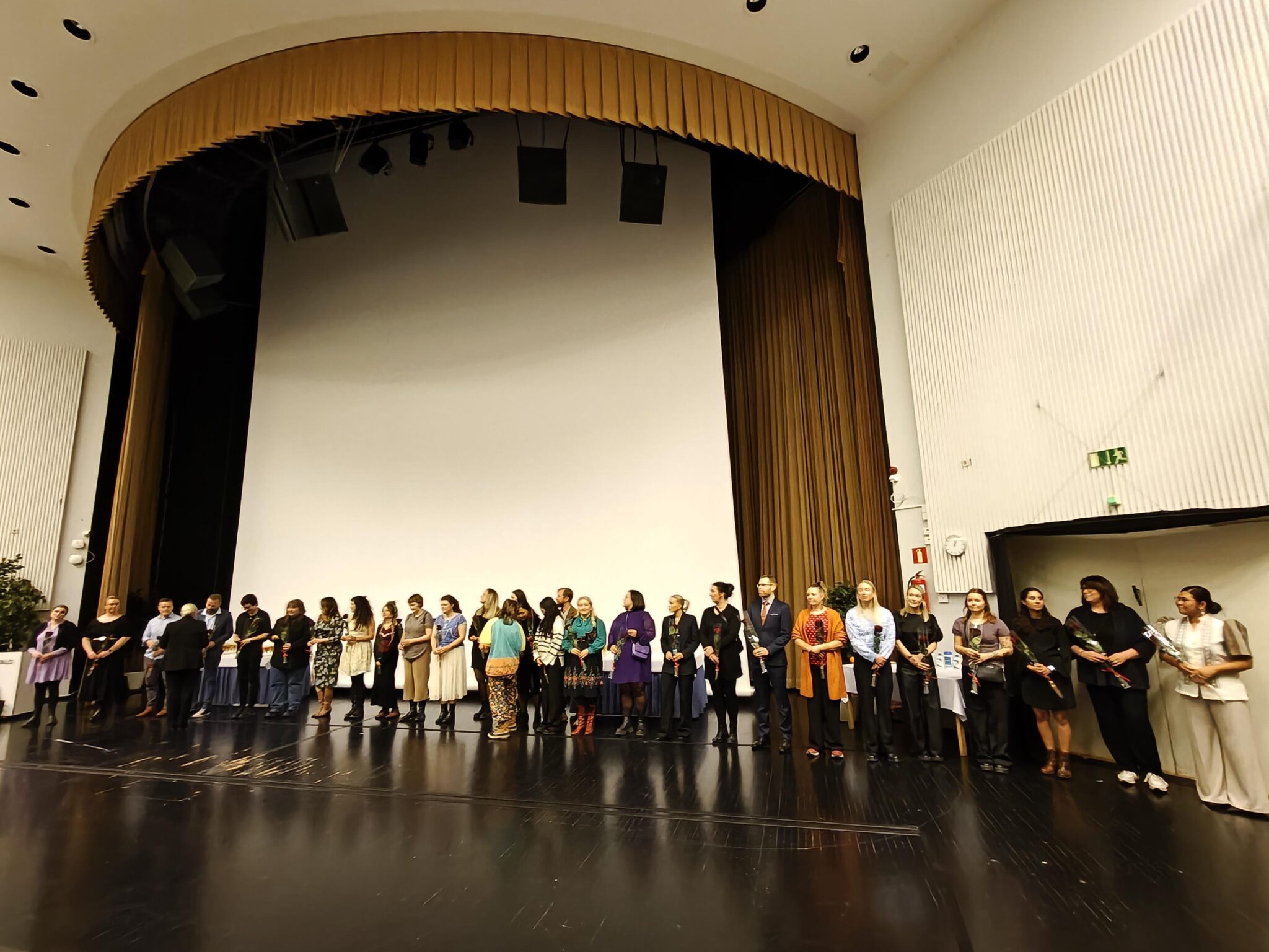 “Suuri joukko aikuisia seisoo rivissä näyttämöllä juhlasalissa. Heillä on yllään erilaisia juhla- ja arkivaatteita, ja useat pitävät kädessään kukkia tai asiakirjoja. Taustalla näkyy korkea valkokangas ja näyttämöverhot, ja tila on valaistu teatterimaisilla valoilla. 