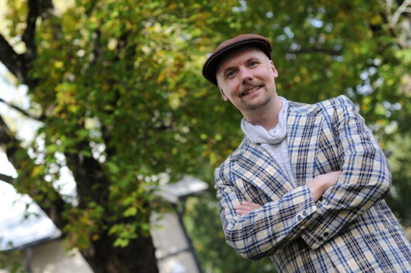 Hattupäinen mies hymyilee kädet puuskassa, yllään kirjava ruudullinen takki ja taustalla puita.