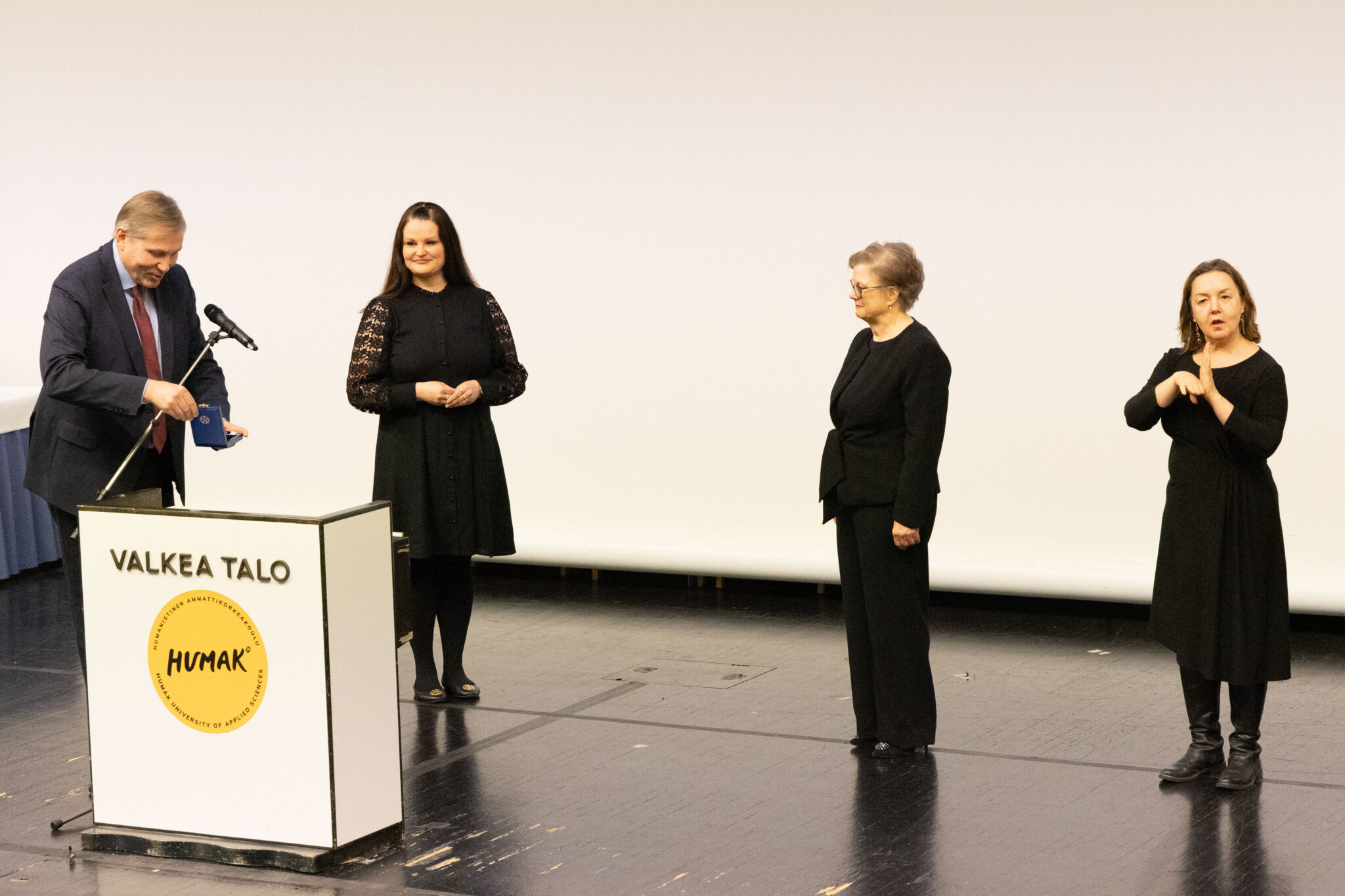 Humakin henkilökuntaa auditorion näyttämöllä. He ovat pukeutuneet tummiin ja juhlavasti. Rehtori-toimitusjohtaja on juuri ojentamassa kunniamerkkiä.