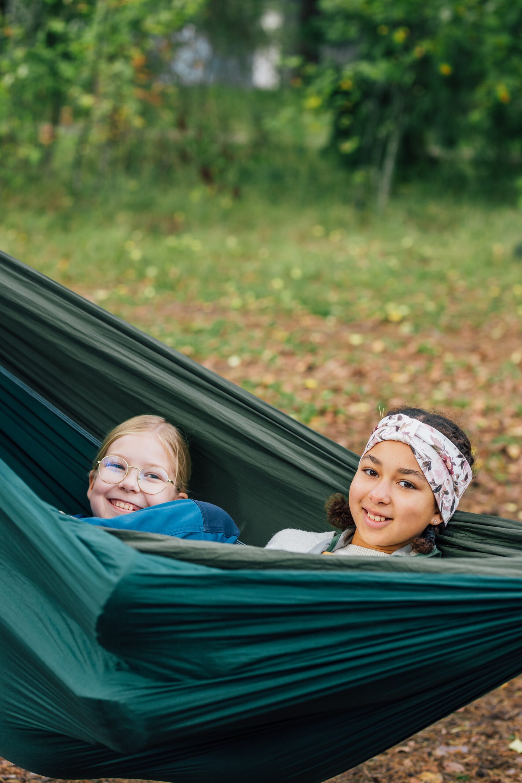 Kaksi henkilöä makaa vierekkäin tummanvihreässä riippumatossa metsäisessä ympäristössä. Riippumatto on levitetty lähelle maata, ja taustalla näkyy lehtipeitteinen maaperä ja vihreää kasvillisuutta.