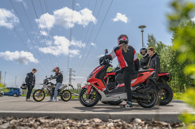 Ryhmä ihmisiä seisoo moottoripyörien ja skootterin ympärillä pysäköintialueella; useilla on ajovarusteet ja kypärät, taustalla sininen taivas, pilviä ja sähkölinjoja.