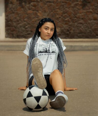 Henkilö istuu kadulla, jalat eteen ojennettuina. Toisen jalan pohja osoittaa kameraan ja jalan alla on jalkapallo. Henkilöllä on tummat pitkät, vaaleiksi taittuvat letit ja vaalea T-paita, jossa on painatusta. Taustalla näkyy kivimuuri ja vihreää kasvillisuutta.