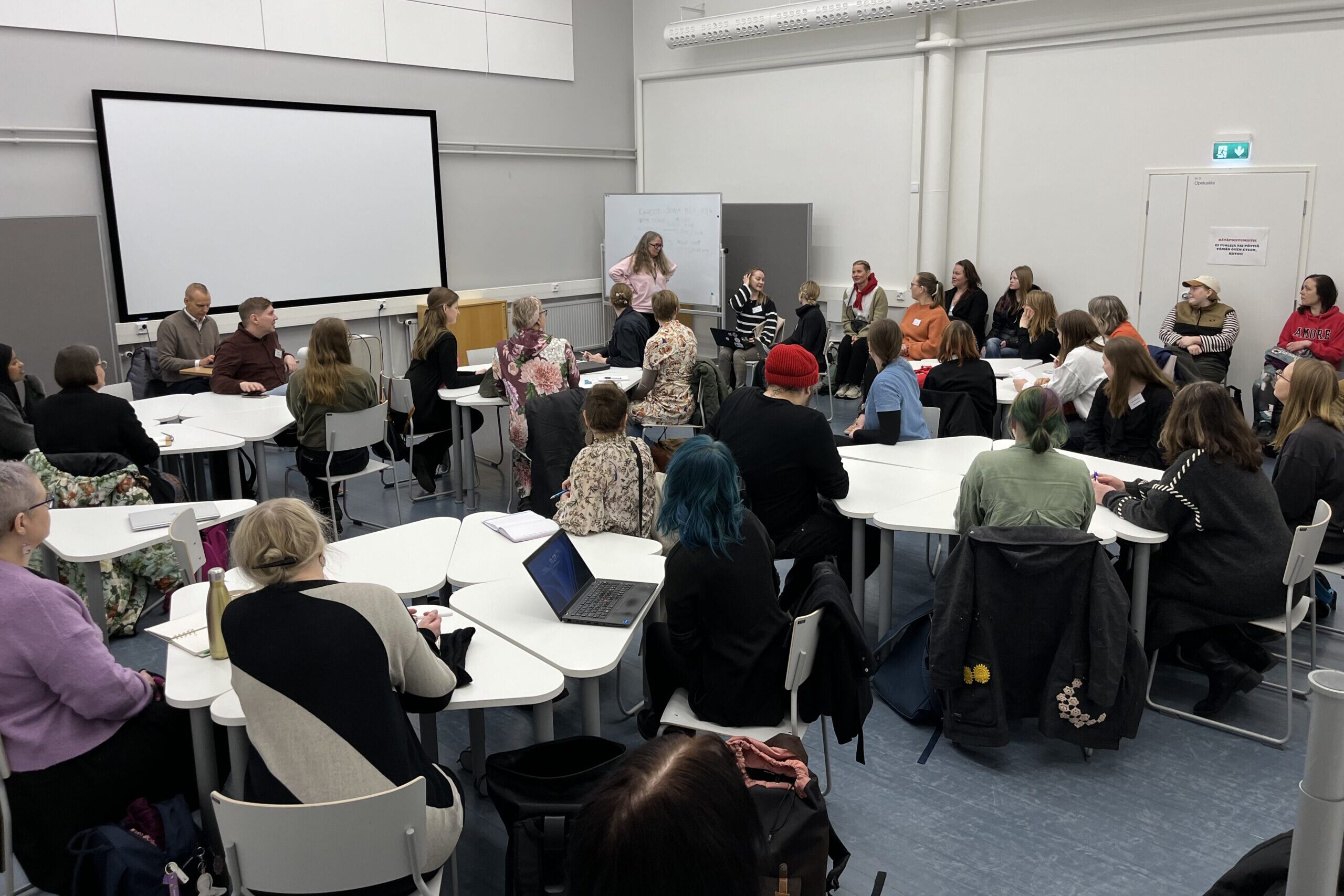 Kuva luokkahuoneesta, jossa ihmiset istuvat pöydissä käymässä ryhmäkeskustelua; pöydillä tietokoneita ja muistiinpanovälineitä.