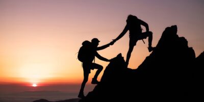 Kuvituskuva, jossa kaksi hahmoa kipuaa kallioista rinnettä ja toinen tukee kädestä vetäen toista kiipeämisessä. Taustalla auringonlasku.