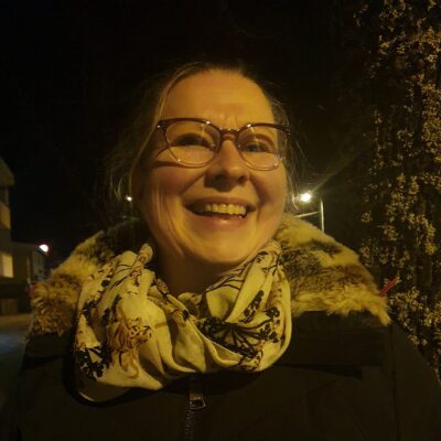 Hymyilevä nainen talvisessa illassa. Naisella on yllään talvitakki ja kaulassaan muhkea huivi.