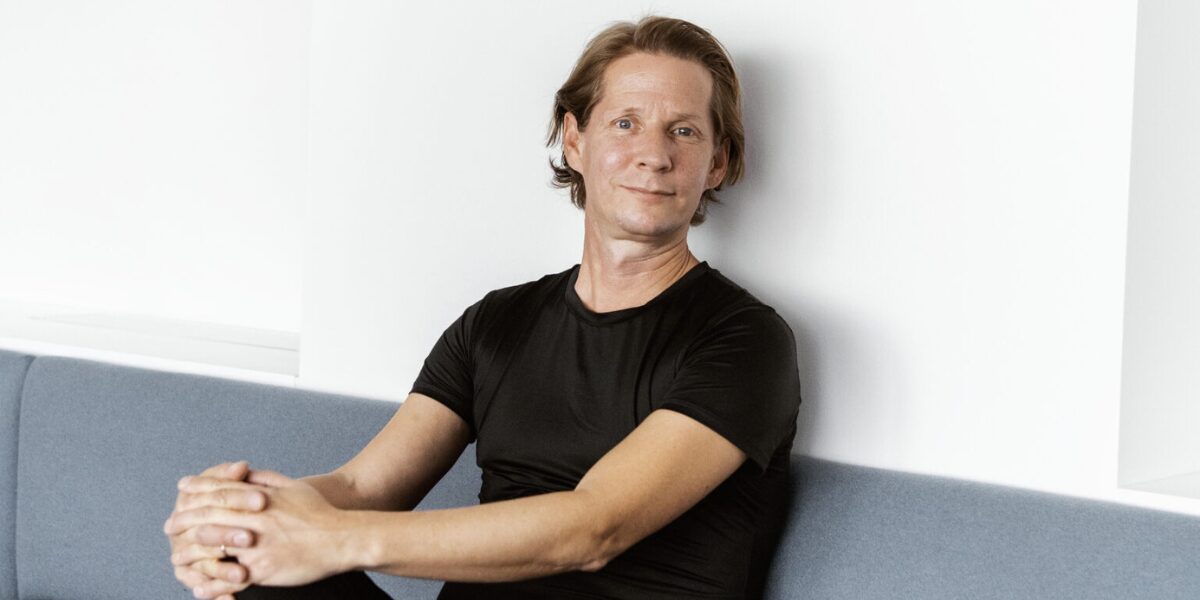 Henkilö istuu kädet sylissä vaaleansinisen sohvan reunalla valkoisen seinän edessä, pukeutuneena mustaan t‑paitaan ja ilmeeltään rentona.