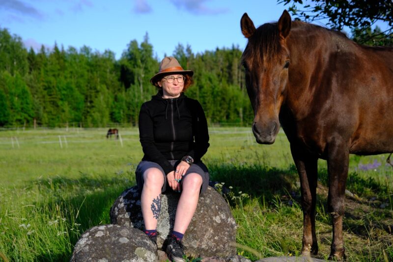 Henkilö istuu kivellä aurinkoisella niityllä mustassa liivissä ja hatussa, sääressä näkyvä tatuointi, ja iso ruskea hevonen seisoo vieressä, taustalla metsää ja laiduntavia hevosia.