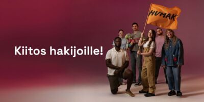 Ryhmakuva iloisesta monimuotoisesta ihmisryhmästä, osa seisoo ja osa polvistuneena, he pitävät oranssia HUMAK-lippua, taustalla punertava liukuväri ja valkoinen teksti "Kiitos hakijoille!"