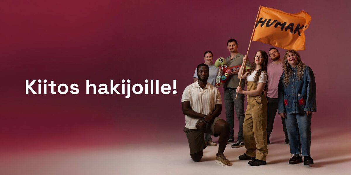 Ryhmakuva iloisesta monimuotoisesta ihmisryhmästä, osa seisoo ja osa polvistuneena, he pitävät oranssia HUMAK-lippua, taustalla punertava liukuväri ja valkoinen teksti "Kiitos hakijoille!"