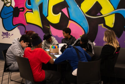 Kuusen nuoren joukko pöydän ääressä piirtämässä, kaksi aikuista ammattilaista heidän kanssaan värikkäässä nuorisotilaympäristössä.