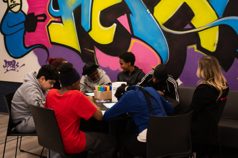 Kuusen nuoren joukko pöydän ääressä piirtämässä, kaksi aikuista ammattilaista heidän kanssaan värikkäässä nuorisotilaympäristössä.